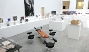 Abgebildet ist die Ausstellung "Werner Ruhnau. Bauen für die offene Gesellschaft" im Baukunstarchiv NRW, die Modelle, Zeichnungen und Fotografie rund um das Werk des Architekten Werner Ruhnau präsentiert. Die Modelle, Fotos, Zeichnungen sind an der Wand und auf Sockeln im Raum verteilt platziert.