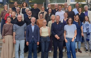 Männern und Frauen stehen verteilt auf Stufen auf Terrasse im Außenbereich und lächeln in die Kamera.