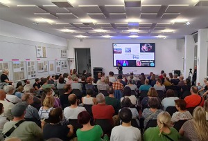 Eine große Menschenmenge sitzt und steht in einem Raum mit weißen Wänden, die teilweise mit Infotafeln behangen sind. Vor der Menschenmenge steht eine Person mit einem Mikrofon in der Hand.