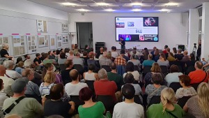 Eine große Menschenmenge sitzt und steht in einem Raum mit weißen Wänden, die teilweise mit Infotafeln behangen sind. Vor der Menschenmenge steht eine Person mit einem Mikrofon in der Hand.