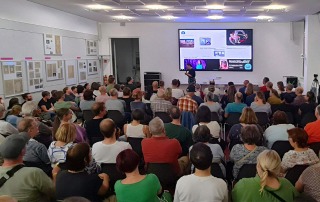 Eine große Menschenmenge sitzt und steht in einem Raum mit weißen Wänden, die teilweise mit Infotafeln behangen sind. Vor der Menschenmenge steht eine Person mit einem Mikrofon in der Hand.