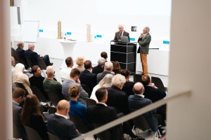 Einige Männer und Frauen sitzen auf Stühlen und schauen zu zwei Männern, die an einem Pult stehen und zur Menge gewandt sind. Die Personen befinden sich in einem Raum mit weißen Wänden.