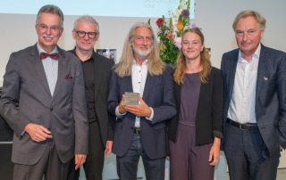 Eine Gruppe Menschen schaut und lächelt in eine Kamera. Ein Mann in der Mitte zeigt eine Medaille, u.a. mit der Aufschrift "Friedrich Halstenberg Medaille". - Foto: Detlef Podehl