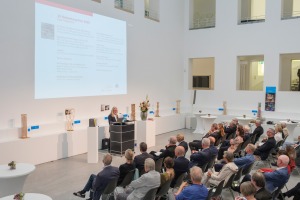 Eine Gruppe Menschen sitzt in einem großen Raum, an einem Pult steht ein Mann und hält einen Vortrag.