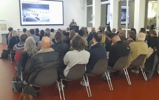 Viele Leute sitzen in einem Raum und schauen auf eine Leinwand, an einem Pult steht ein Mensch und redet zur Personengruppe.