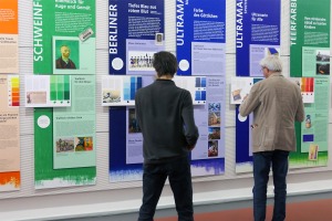 Zwei Männer stehen vor bunten Informationstafeln zum Thema Farben und Pigmente und schauen sich die Informationstafeln an.