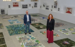 Blick in einen Ausstellungsraum. Auf dem Boden liegen Teppiche mit Gemälden und Fotografien, an der Wand stehen auf Sockeln Fotografien. Zwei Personen stehen im Raum und schauen in die Kamera. - Foto: Detlef Podehl