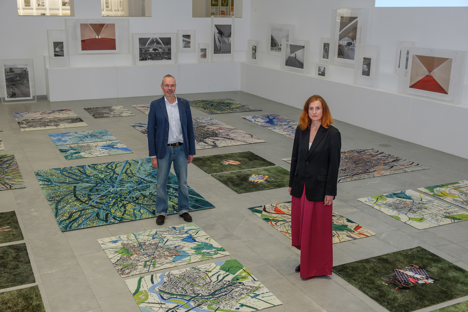 Blick in einen Ausstellungsraum. Auf dem Boden liegen Teppiche mit Gemälden und Fotografien, an der Wand stehen auf Sockeln Fotografien. Zwei Personen stehen im Raum und schauen in die Kamera. - Foto: Detlef Podehl