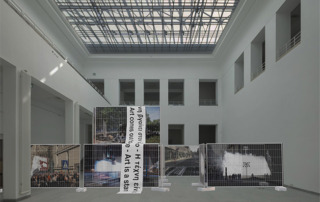 Grafik/Collage: Ein Ausstellungsraum, in der Mitte sind Bauzäune mit Bannern. - Bildcredit: Simulation public preposition: Baukunstarchiv NRW © Studio Mischa Kuball