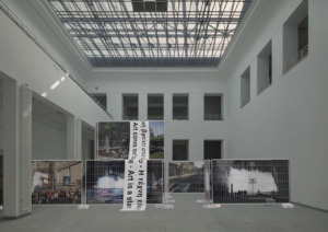 Grafik/Collage: Ein Ausstellungsraum, in der Mitte sind Bauzäune mit Bannern. - Bildcredit: Simulation public preposition: Baukunstarchiv NRW © Studio Mischa Kuball