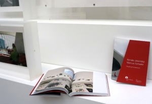 Auf weißen Sockeln sind gerahmte Fotografien und zwei Bücher positioniert. Ein Buch ist geschlossen, ein zweites aufgeklappt positioniert.