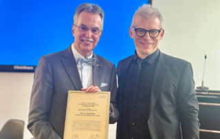Zwei Männer, die lächeln, einer von beiden hält eine gerahmte Urkunde in der Hand.