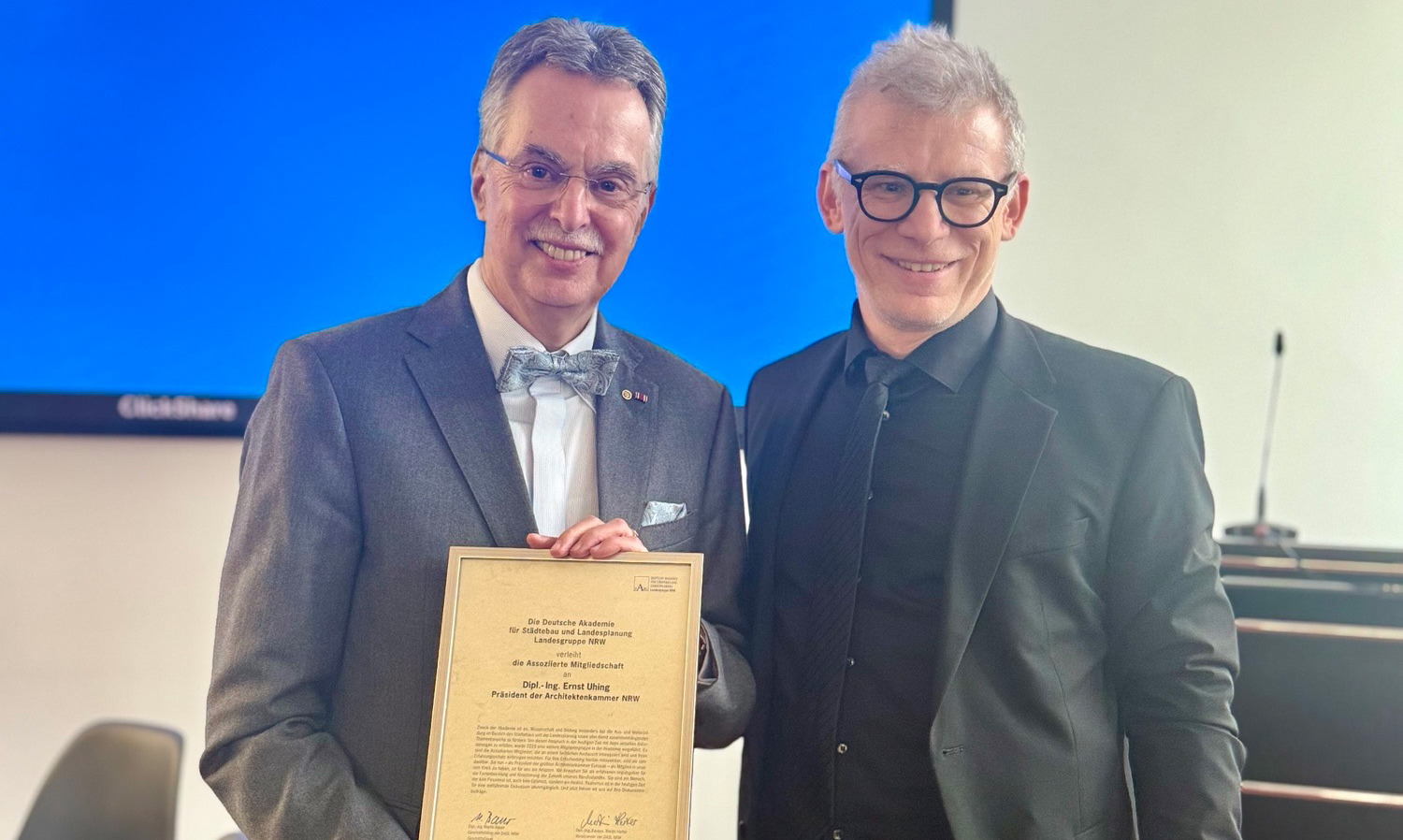 Zwei Männer, die lächeln, einer von beiden hält eine gerahmte Urkunde in der Hand.