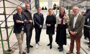 Eine Personengruppe steht beisammen und lächelt in die Kamera. Im Hintergrund unterhalten sich Menschen miteinander.