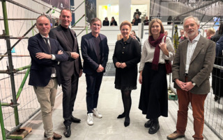 Eine Personengruppe steht beisammen und lächelt in die Kamera. Im Hintergrund unterhalten sich Menschen miteinander.