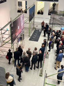 Personen stehen in einem hellen Raum, in dem Bauzäune mit Transparenten hängen. Sie schauen größtenteils zu einem Pult.