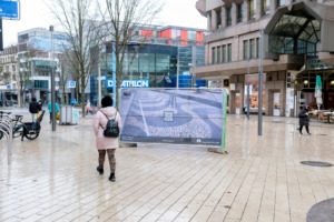 Bauzäune mit Plakaten stehen in einer Innenstadt herum, Personen mit Mützen und Kapuzen laufen an den Bauzäunen vorbei.