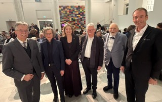 Sechs Personen stehen vor einer Menschengruppen und lächeln in die Kamera.