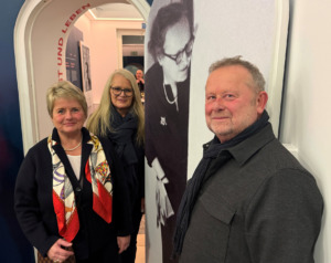 Drei Personen lächeln in die Kamera und stehen an einer Tür mit einem Poster, das eine Frau zeigt.