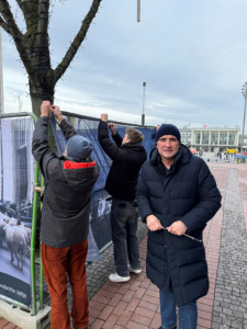 Personen bauen an einem Bauzaun im Außenraum eine Installation auf und hängen Banner auf. Ein Mann lächelt in die Kamera.