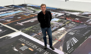 Ein Mann steht in einem Raum und lächelt in die Kamera, Banner liegen auf dem Boden und zeigen Kunstprojekte.