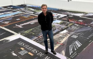 Ein Mann steht in einem Raum und lächelt in die Kamera, Banner liegen auf dem Boden und zeigen Kunstprojekte.