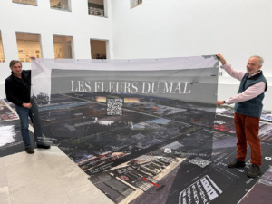 Zwei Männer stehen in einem hellen Raum und halten ein Banner hoch auf dem unter anderem steht 