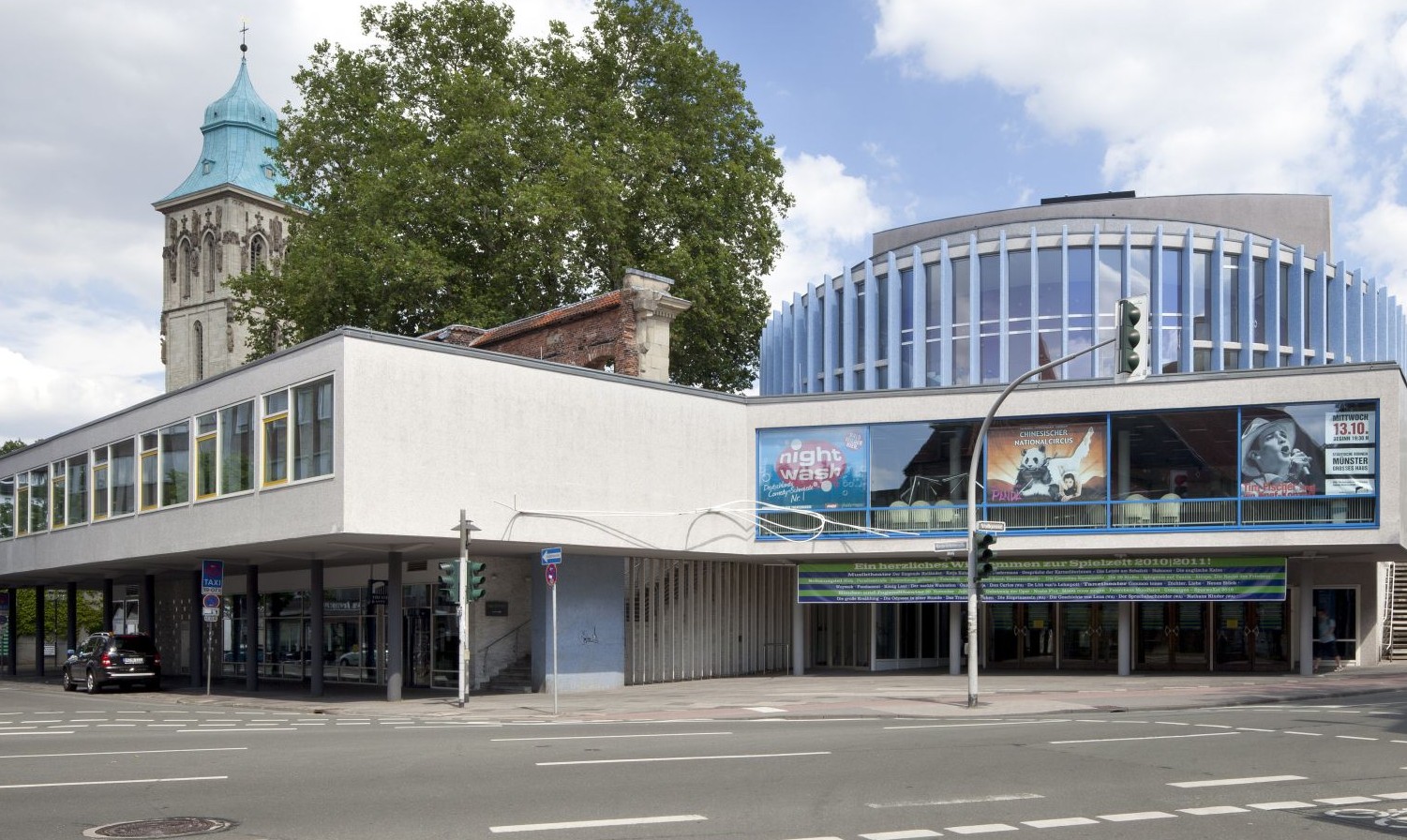Ein Gebäude an einer Straßer gebaut, im Hintergrund sind Bäume und ein blauer Himmel mit Wolken.