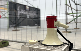 Eine Megafon liegt auf einer Kiste, dahinter sind Bauzäune, teilweise bespannt mit Plakaten.