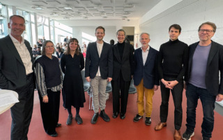 Eine Gruppe an Männern und Frauen steht beieinander. Die Menschen lächeln teilweise in die Kamera.