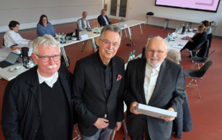 Drei Männer stehen lächeln und stehen beeinander, im Hintergrund sitzen mehrere Personen an einem Tisch.