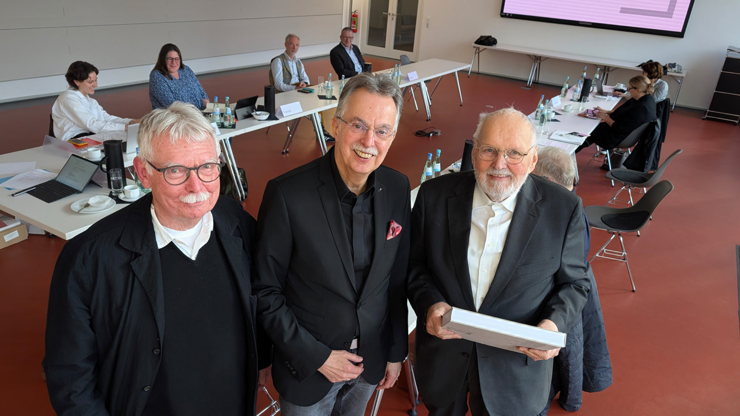 Drei Männer stehen lächeln und stehen beeinander, im Hintergrund sitzen mehrere Personen an einem Tisch.