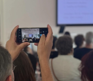 Menschen sitzen in einem Raum, eine Person fotografiert die Situation mit einem Smartphone.